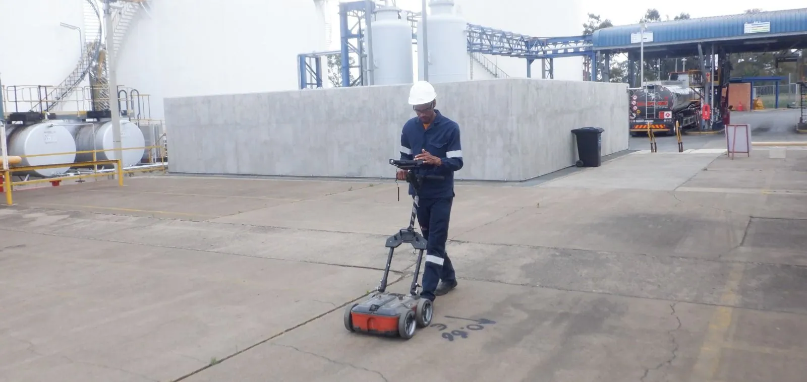 An HD GPR technician scanning a concrete yard with a ground penetrating radar cart, with storage tanks behind